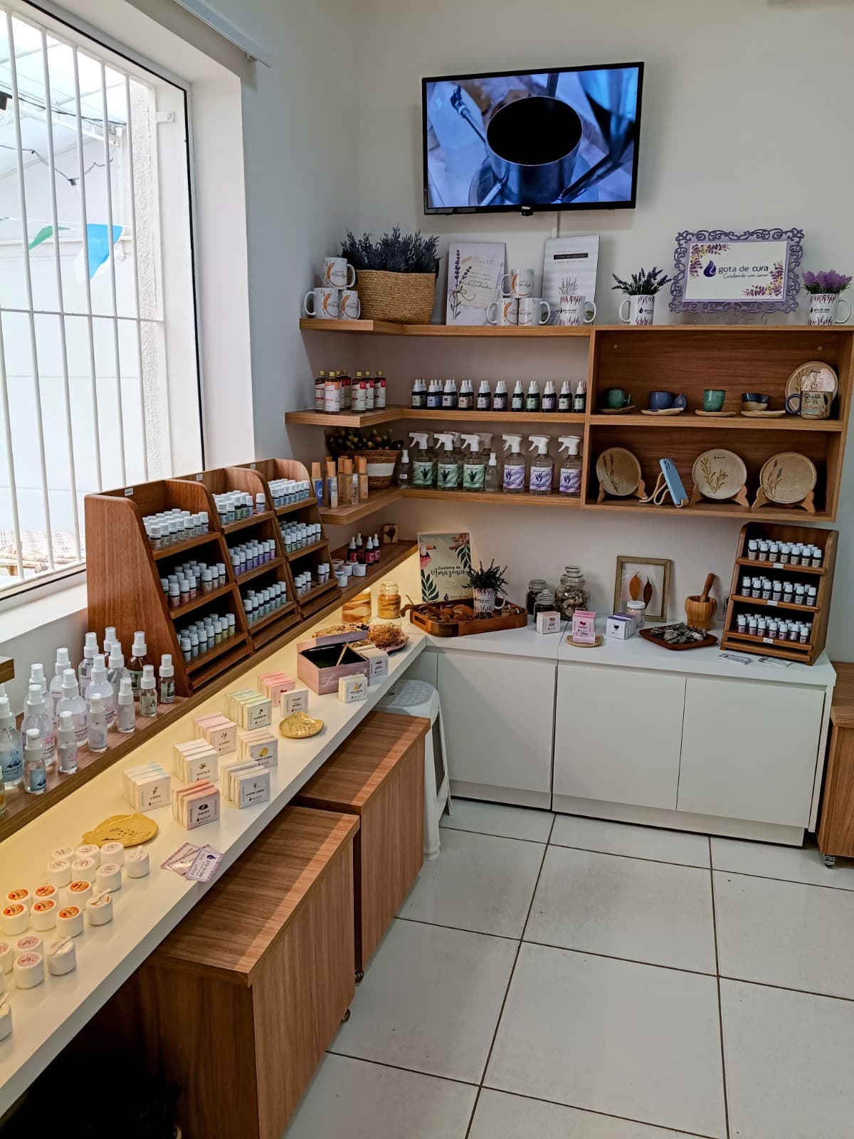 O interior da loja da Gota de Cura: prateleiras de madeira com sprays, óleos essenciais e sabonetes, o quadro da marca na parede e a luz entrando pela janela.